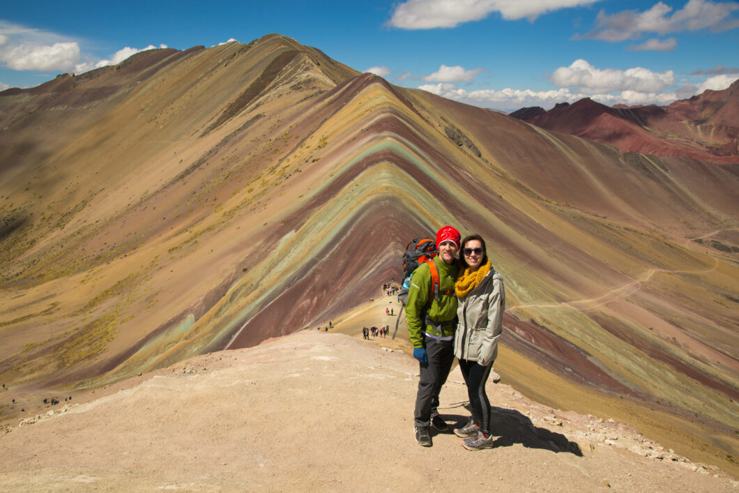 A couple standing and smiling towards the camera on top of Rainbow mountain in Peru.