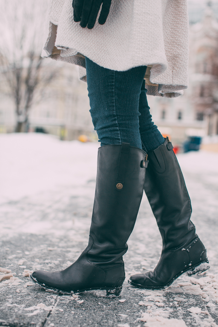 A closeup of a women's legs and feet. She's standing on a snowy sidewalk, wearing knee-high black leather boots over skinny jeans.