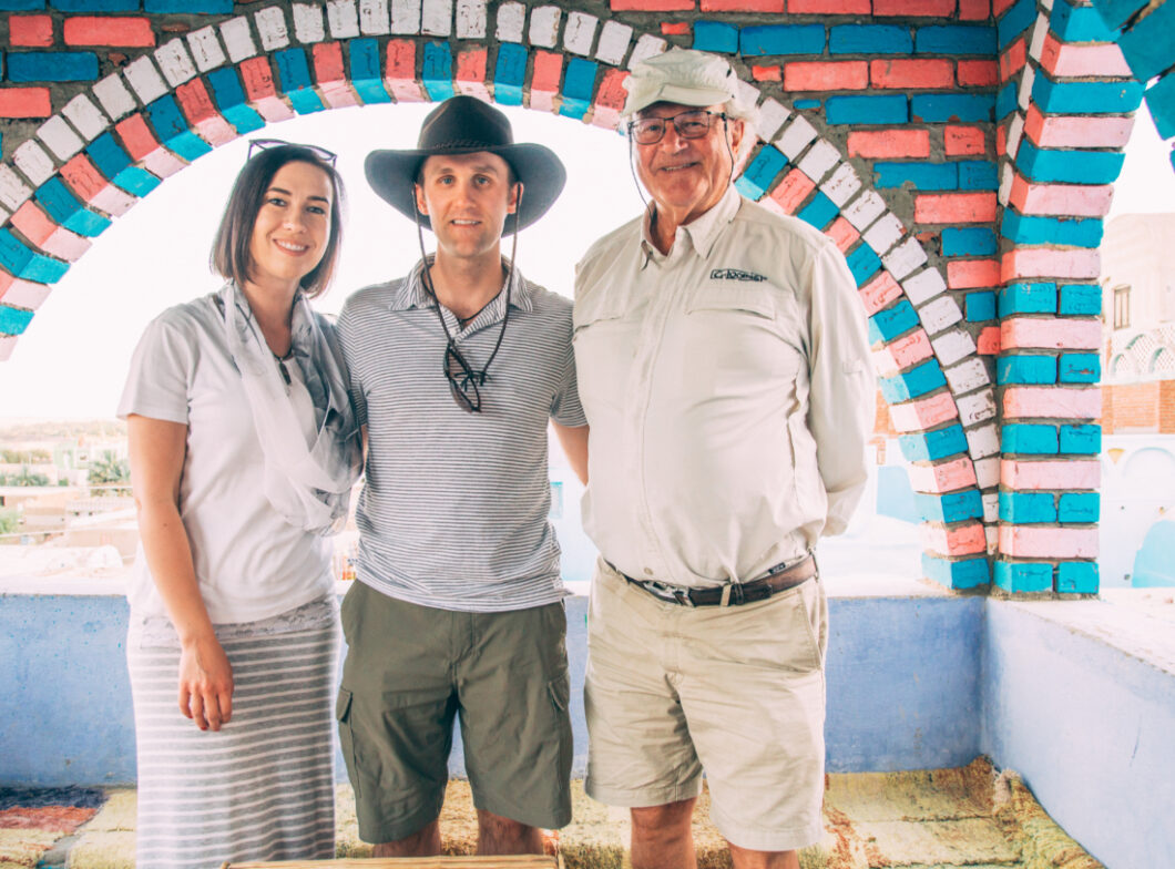 Three people pose together for a photo. The young woman wears a skirt and short-sleeved shirt with a scarf around her neck. The man in the middle wears a striped collard shirt, dark khaki shorts, and a wide-brimmed hat, and the older man on the end wears a long sleeved white shirt and light khaki shorts.