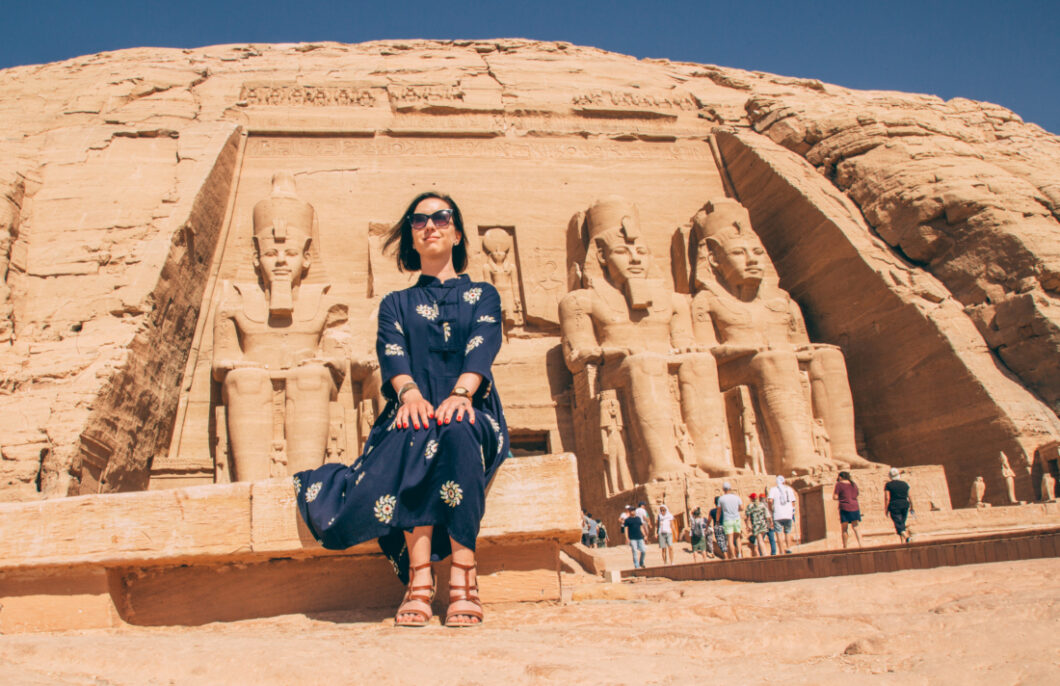 A woman sits on a bench in front of the entrance of the Ramses II temple in Abu Simbel. She poses with her hangs on her knees, similar to the giant stone Ramses statues behind her. Tourists in the background line up to enter the temple.