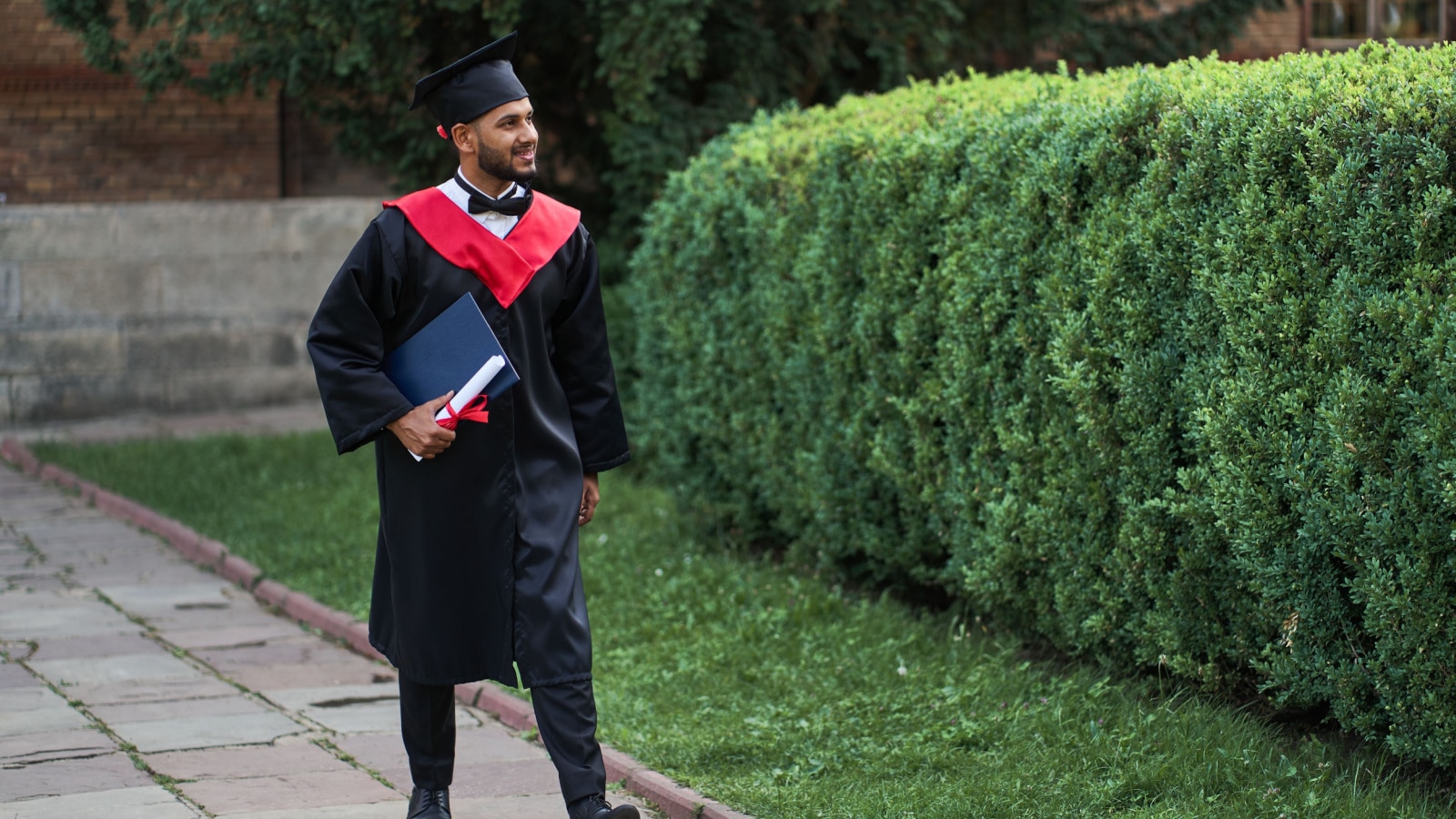Học sinh đẹp trai người Ấn Độ đi bộ trên khuôn viên trường đại học trong bộ áo choàng tốt nghiệp và bằng cấp, không gian để sao chép