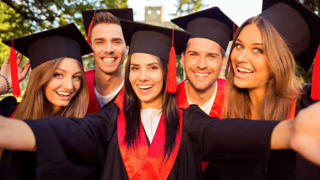 A group of five college graduates wearing grad caps and gowns, smiling at the camera while taking a selfie.