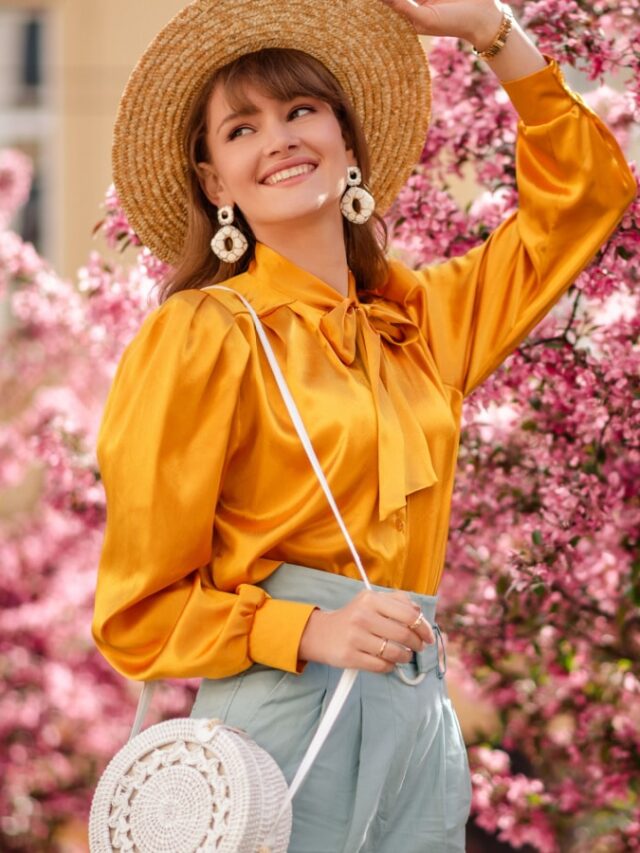 Outdoor lifestyle portrait of elegant happy smiling woman wearing trendy straw hat, yellow satin blouse, blue trousers, with shoulder wicker round bag, walking in street near pink spring blossom trees