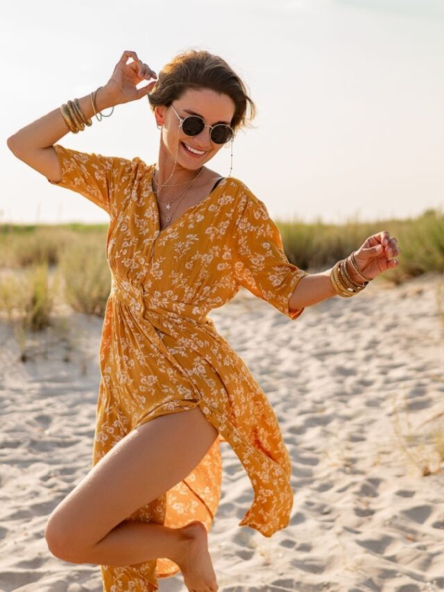 stylish attractive slim smiling woman on beach in summer style fashion trend outfit carefree and happy, feeling freedom, wearing yellow printed dress boho style chic and sunglasses