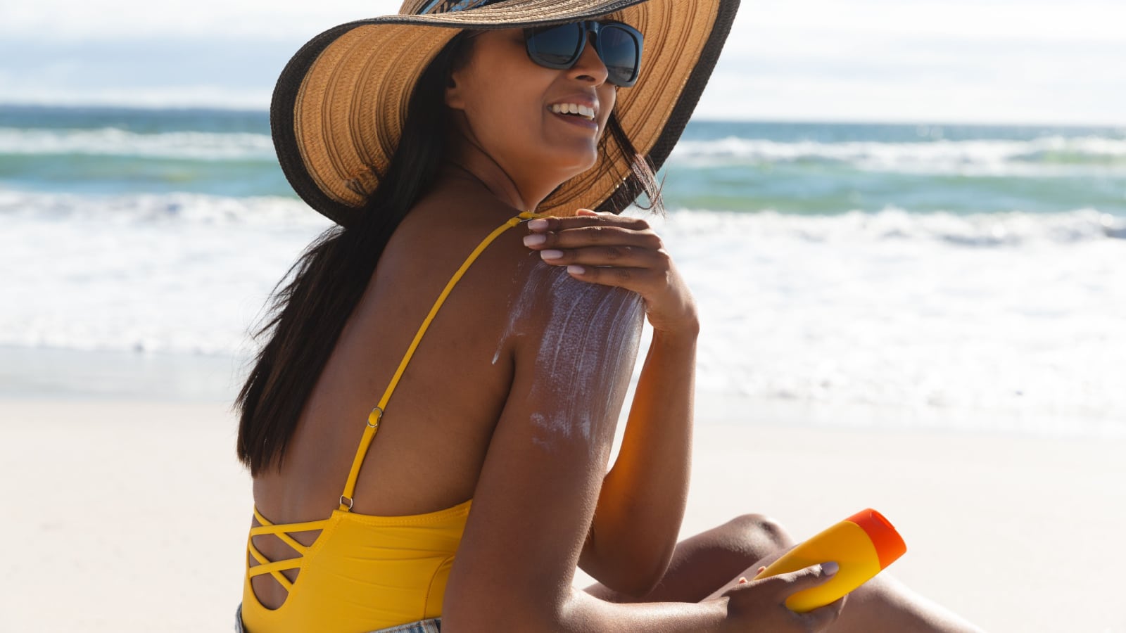 Smiling mixed race woman on beach holiday using sunscreen cream. healthy outdoor leisure time by the sea.