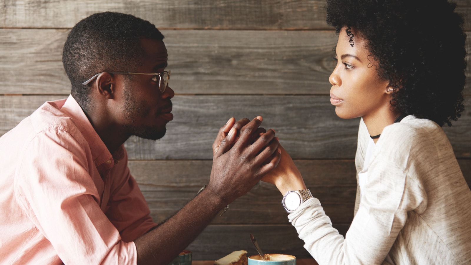 African American young couple sitting at a cafe table: black man in glasses holding her beautiful wife's hands, begging her to forgive him, apologizing for an affair, looking with guilty expression