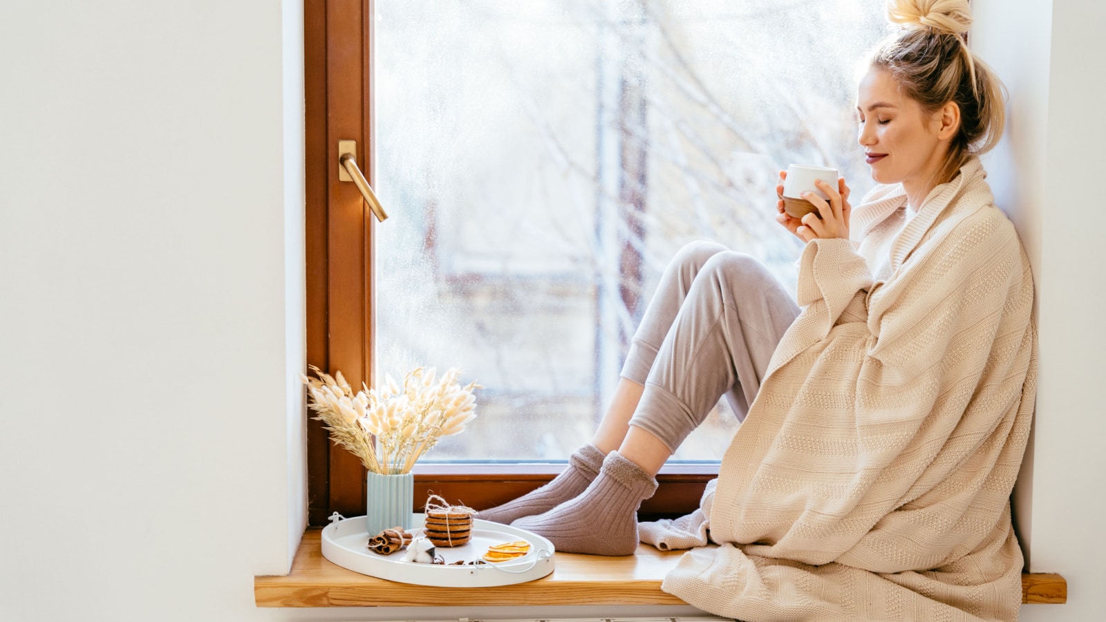 Happy blond woman with bunch hairstyle warming and cover knitted plaid enjoying in her coffee time by the window in cold winter day. Peace of mind and mental health.