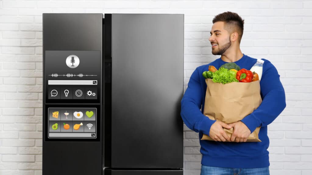 Young man with bag of groceries near smart refrigerator indoors