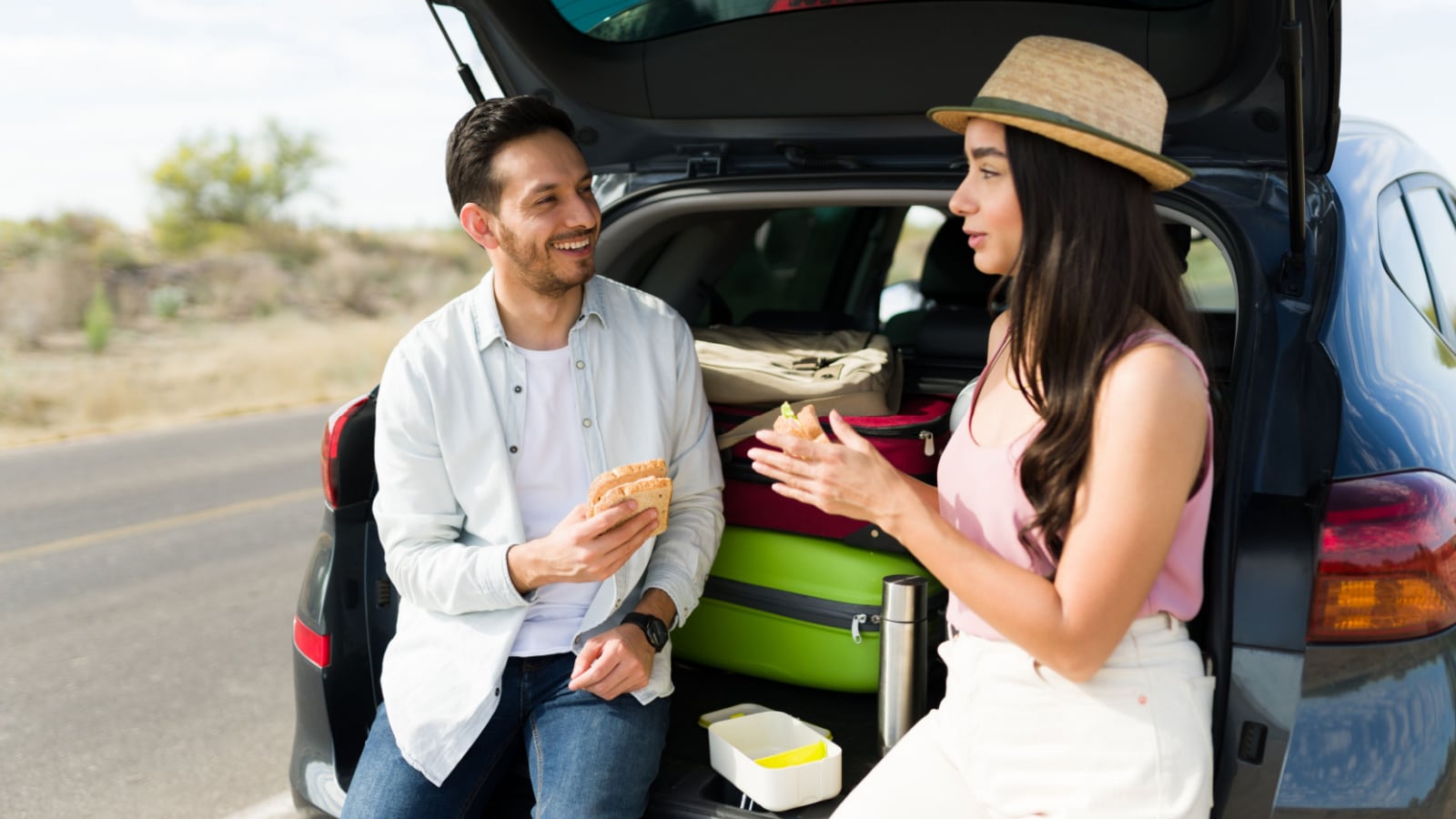 Attractive young man and woman eating a sandwich while resting on the car trunk during a weekend trip together