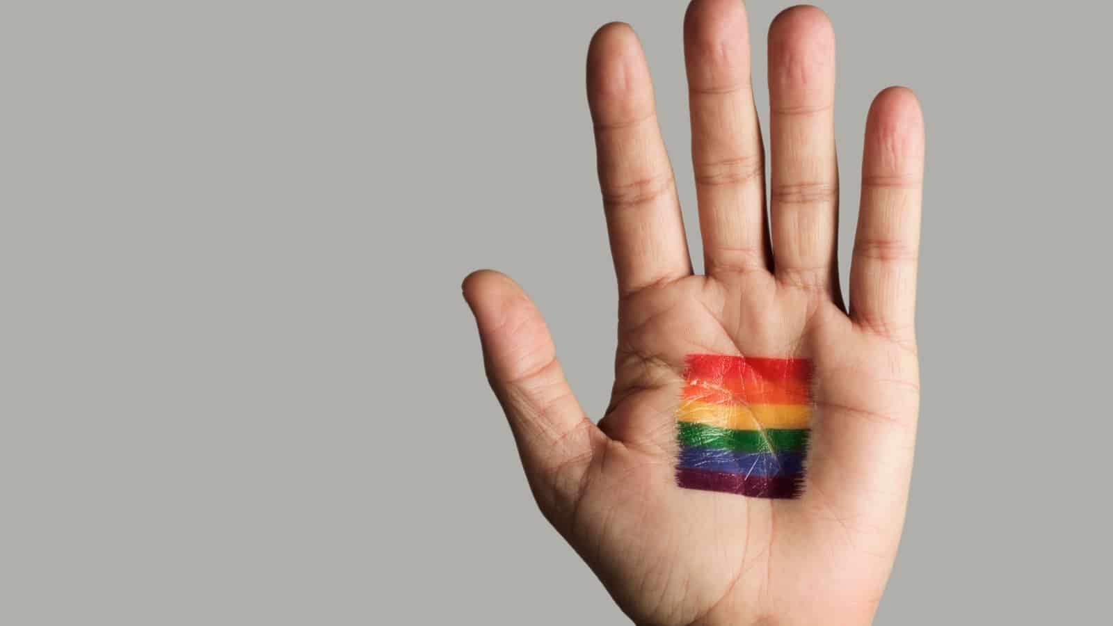 closeup of a rainbow flag in the palm of the hand of a young caucasian person, against an off-white background with some blank space on the left