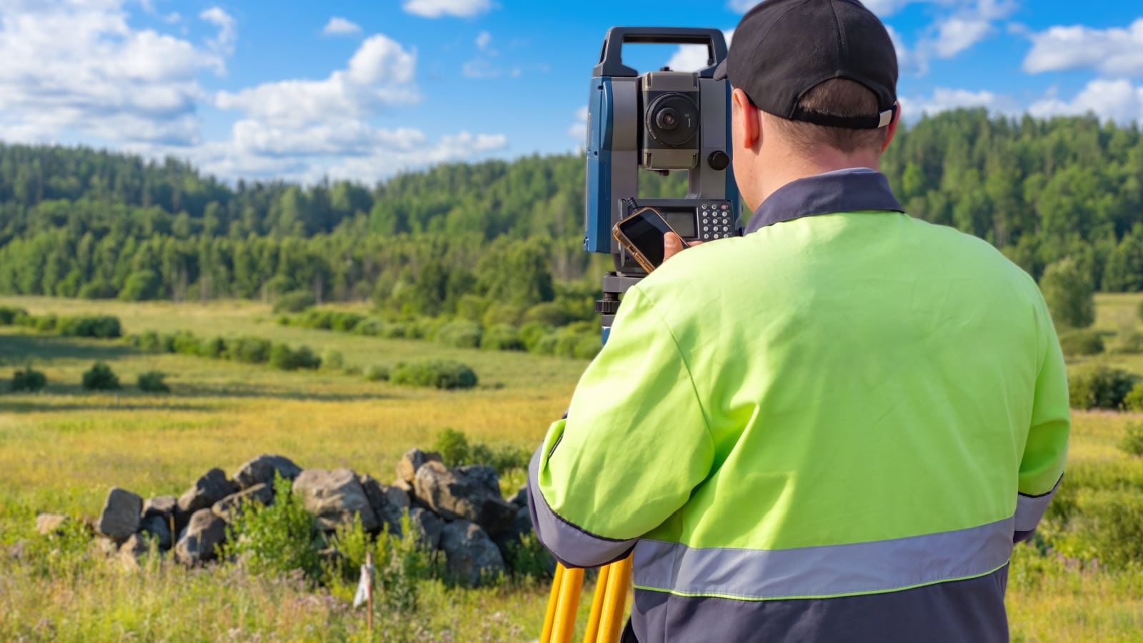 Man surveyor with his back to camera. Specialist in geodesy near forest. Builder surveyor measures plot of land. Man worker with optical theodolite. Surveyor inspects construction site.