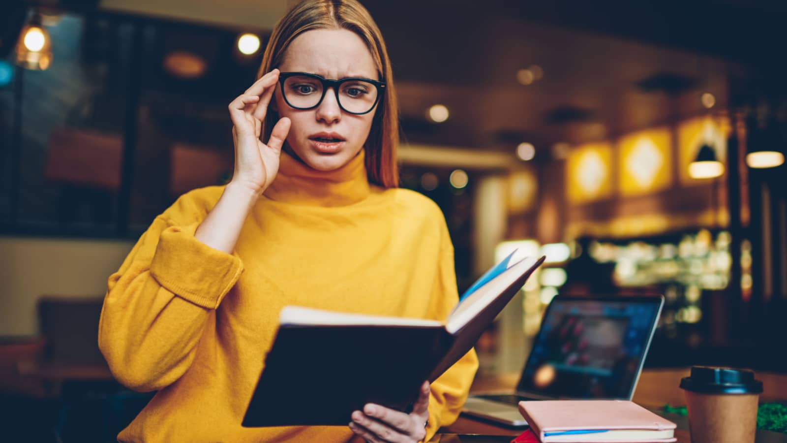 Young attractive emotional female person in cool spectacles surprised while reading popular novel standing in coffee shop.Pretty hipster girl felling scary during checking information from notepad