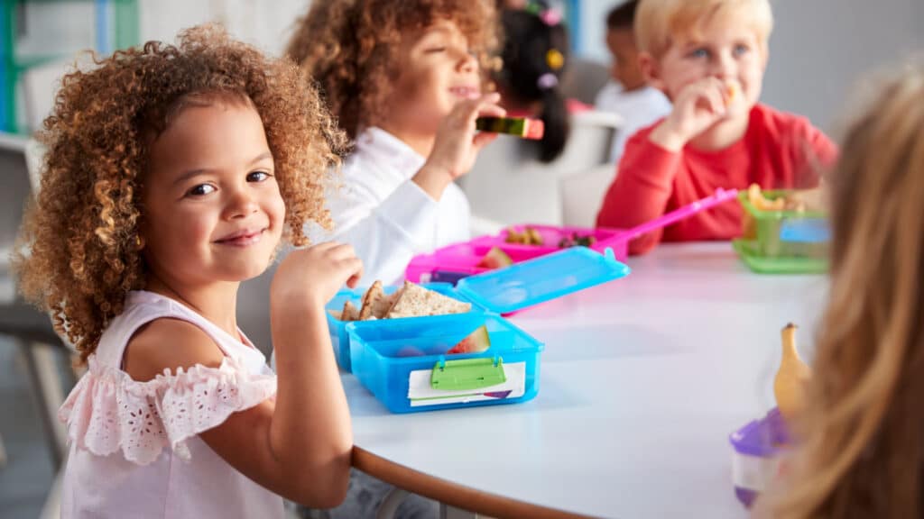 Close up of smiling young children sitting at a table eating their packed lunches together at infant school, girl smiling to camera, selective focus