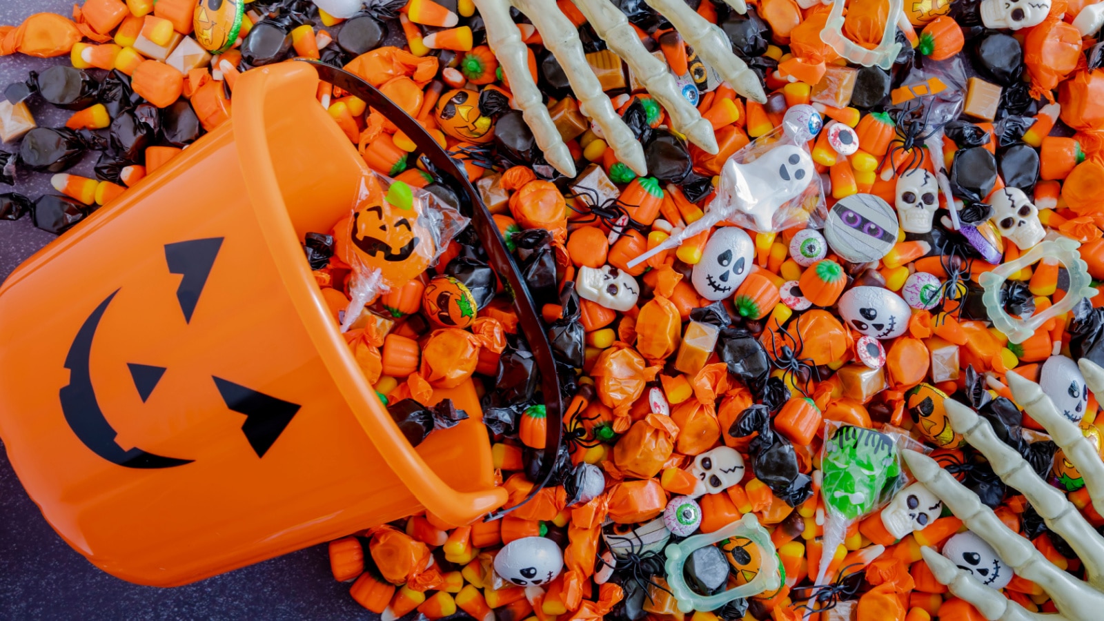 Orange pumpkin pail spilling Halloween candy on black stone surface with skeleton hands grabbing candy