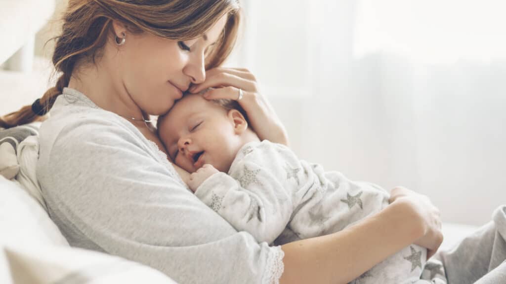 Loving mom carying of her newborn baby at home. Bright portrait of happy mum holding sleeping infant child on hands. Mother hugging her little 2 months old son.