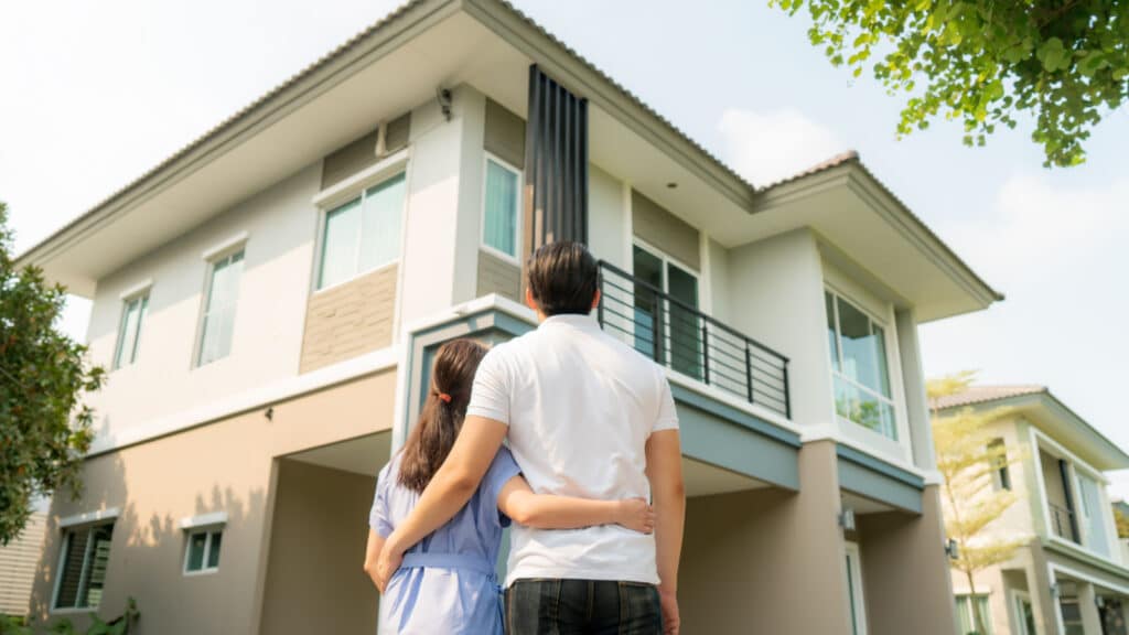 Back portrait of Asian young couple standing and hugging together looking happy in front of their new house to start new life. Family, age, home, real estate and people concept.