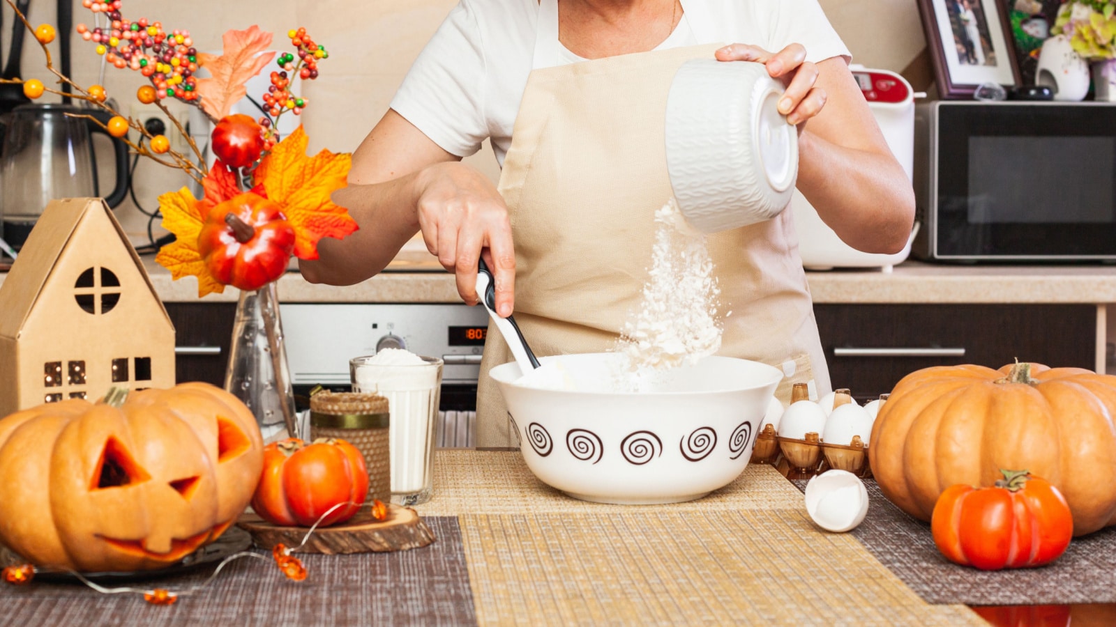 A woman pours flour from a glass into cookie dough for Halloween in the kitchen with an autumn decor. Cozy home. Making cookies for Halloween.