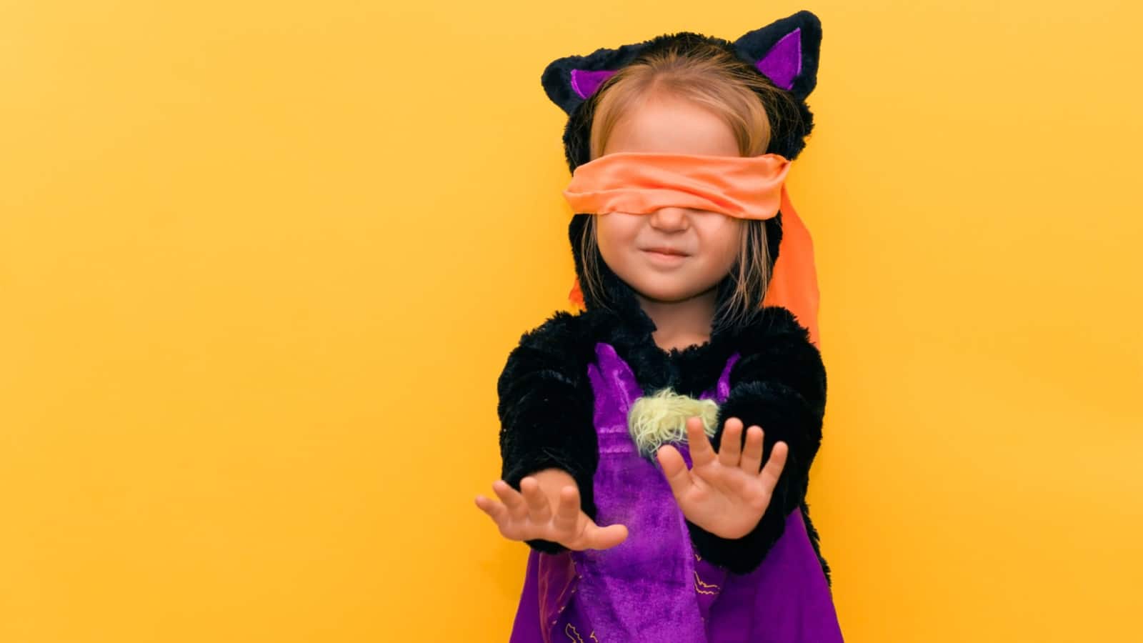 Halloween games for young children. A child in a black purple bat suit blindfolded looking for others. Portrait on a yellow-orange background