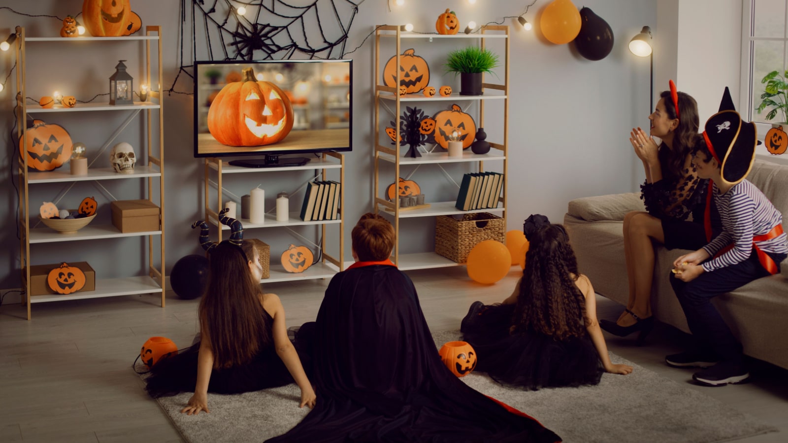 Kids in spooky costumes of witches, monsters and pirates watching children's Halloween movie sitting on floor and sofa in interior decorated with orange and black pumpkins at fancy dress party at home