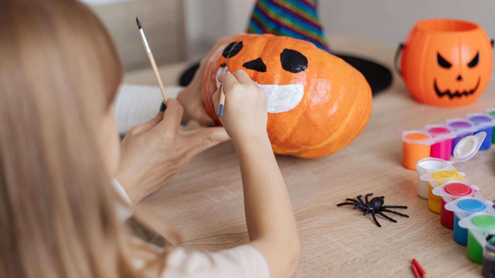 child with mother painting Halloween pumpkins together at home, happy family mother and kid preparing handmade decorations for Saints Day party