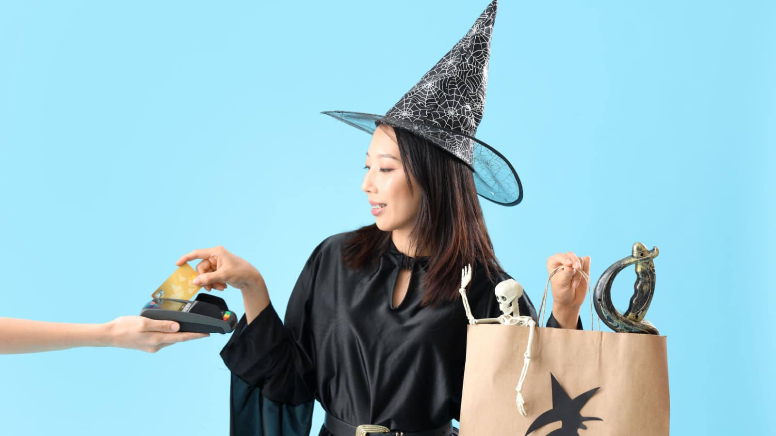 Young Asian woman dressed for Halloween as witch with credit card and shopping bag paying on blue background