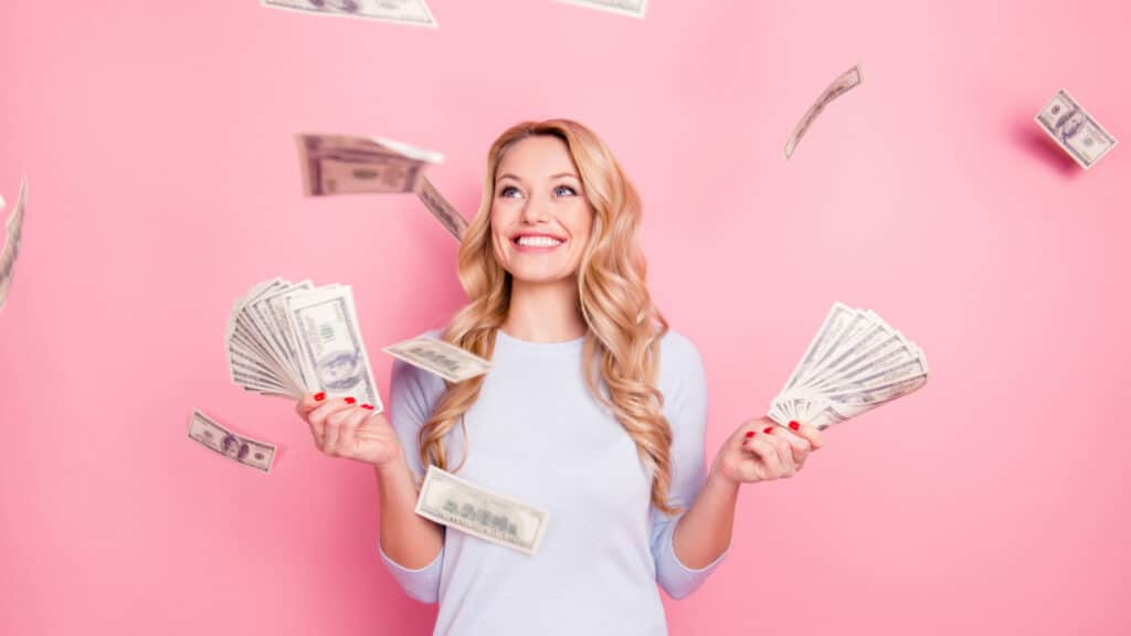 Portrait of pretty charming positive cute successful lucky cheerful girl standing under shower from money having a lot of money in hands isolated on pink background