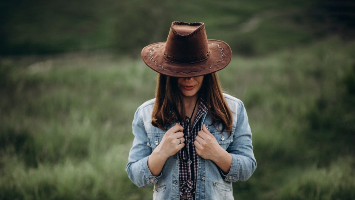 Mysterious girl hides her face behind a cowboy hat. Girl alone in the field in the evening holds on to a denim jacket as if frozen on the head cowboy hat