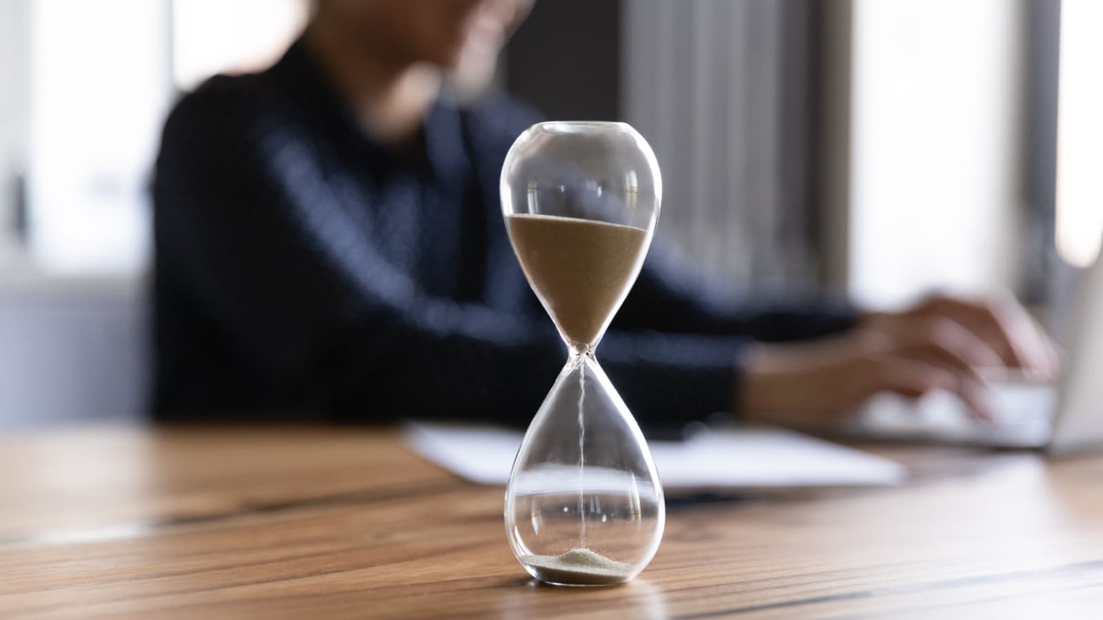 Close up view of hourglass stand on wooden home office measuring time, woman busy using computer. Female employee on background work on laptop, try to meet deadline. Efficiency concept.