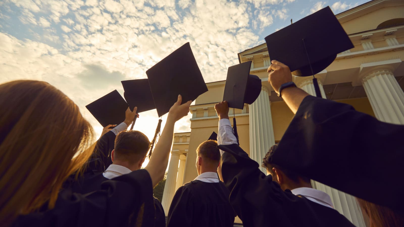 Graduation ceremony event celebration. Diverse student wearing gown mantles standing in group holding raised hat rear view from bottom. Shot over sunset and university high school building