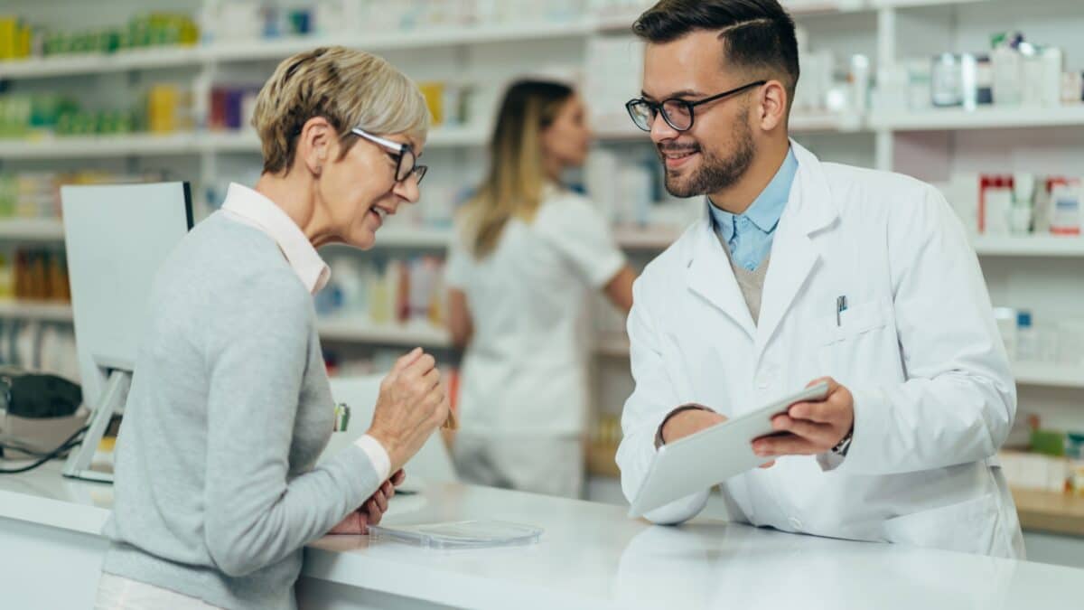 Young male pharmacist giving prescription medications to senior female customer in a pharmacy with female pharmacist in the background