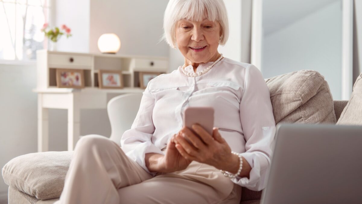 Keeping in touch. Pleasant elderly woman sitting on the couch in the living room and sending text messages to her children while smiling