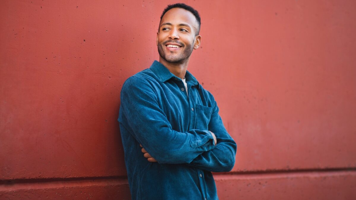 Happy young man standing with arms crossed against maroon wall and looking away