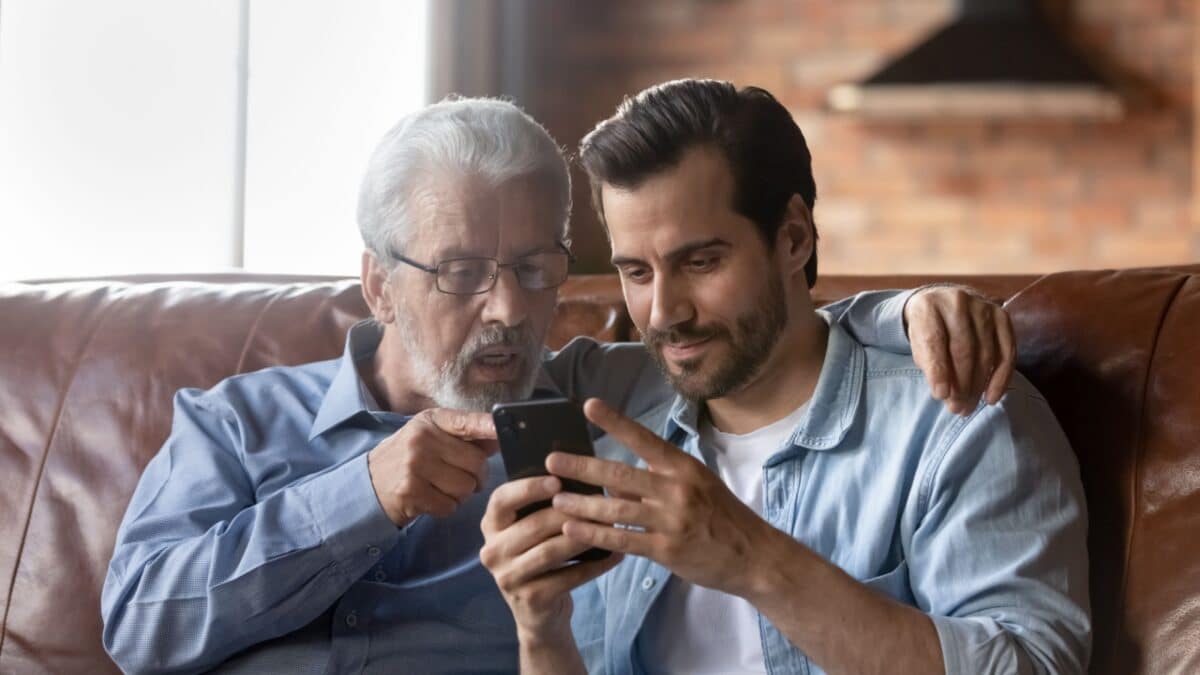 Grown son teaching elderly 70s dad to use mobile app on smartphone, showing family pictures, explaining online payment service. Two family generations men resting on couch, using cellphone together