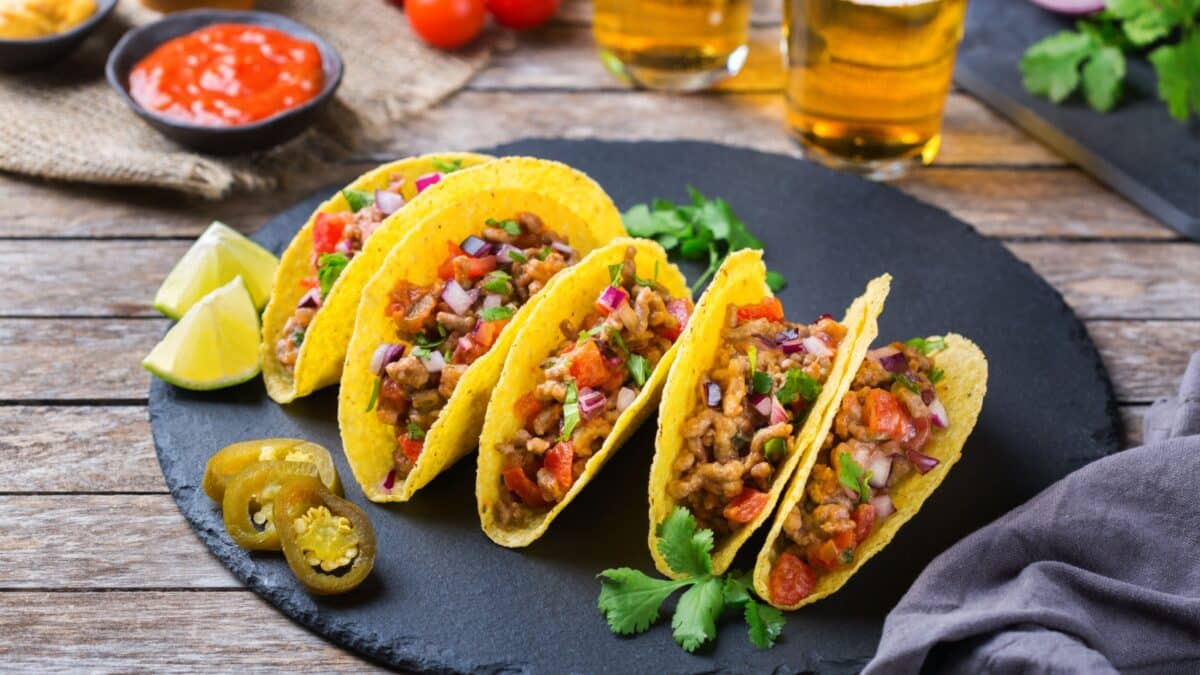 Taco shell ellow corn tortilla chips nachos with ground beef, mince, guacamole, red hot jalapeno chili salsa and cheese sauce with tequila or beer on a table.