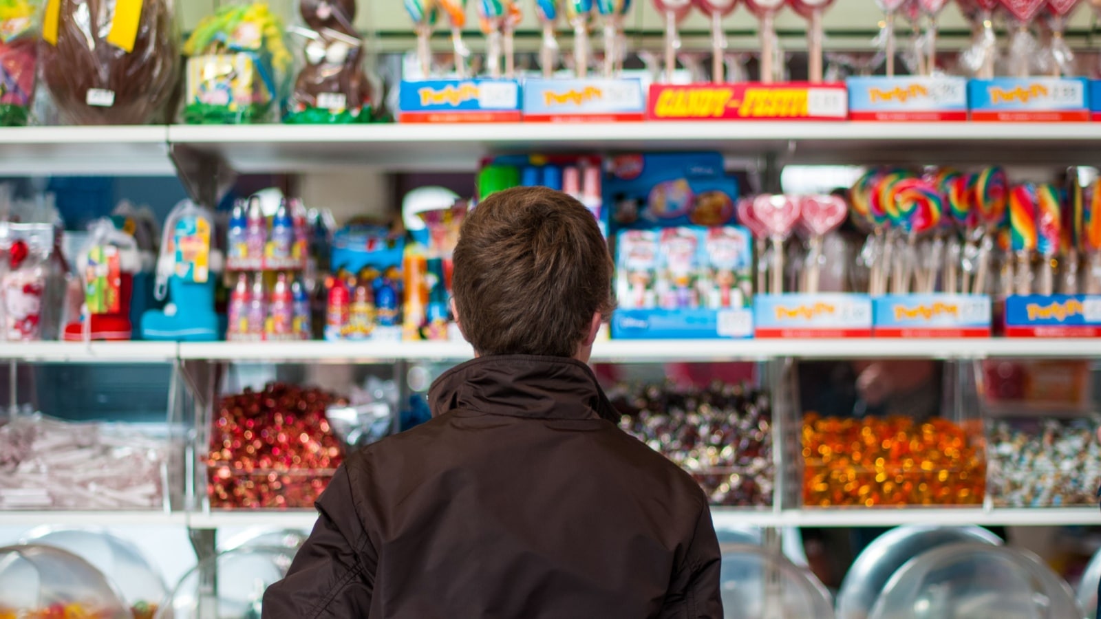 A young person with brown hair wearing a brown casual jacket seen from behind is standing in front of colourful shelves filled with sweets and candy in a store.