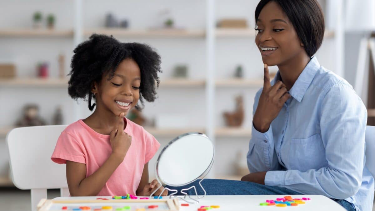 Young black woman speech pathologist exercising with adorable little girl with language difficulties, working on word sounds, kid looking at mirror, touching chin and smiling, clinic interior