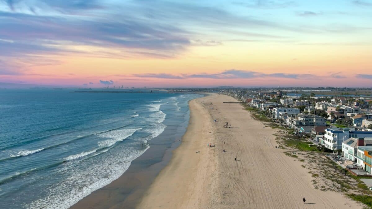 aerial shot of the coastline with blue ocean water and homes along the sand on the beach, cars driving on the street and blue sky with clouds in Huntington Beach California USA