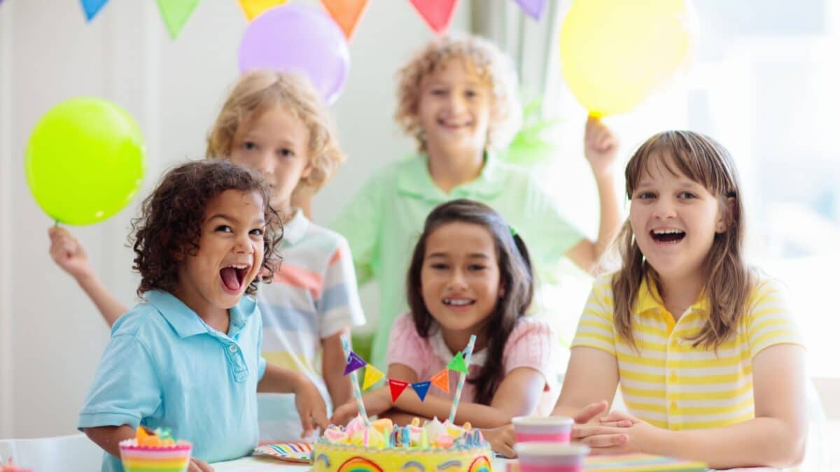 Kids birthday party. Children celebrate with colorful cake and gifts. Little curly boy blowing candles and opening birthday presents. Friends play with rainbow confetti. Party home decoration.