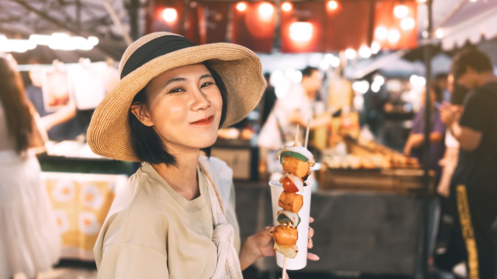 Poeple travel and eating street food concept. Happy young adult asian foodie woman holding bbq grilled skewers at outdoor vendor night market.