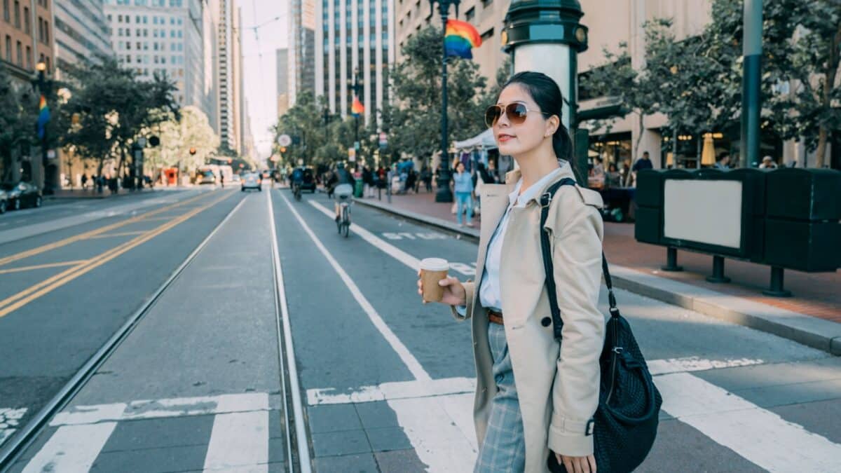 beautiful carefree asian woman worker cross zebra road with paper cup of beverage in busy city. Simple flag LGBT hanging on street lamp in san francisco usa. elegant urban girl in sunglasses and bag