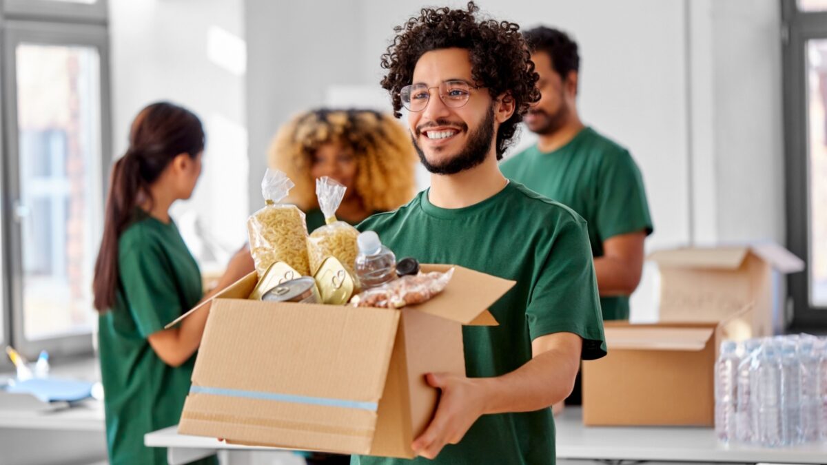 charity, donation and volunteering concept - happy smiling male volunteer with food in box and international group of people at distribution or refugee assistance center