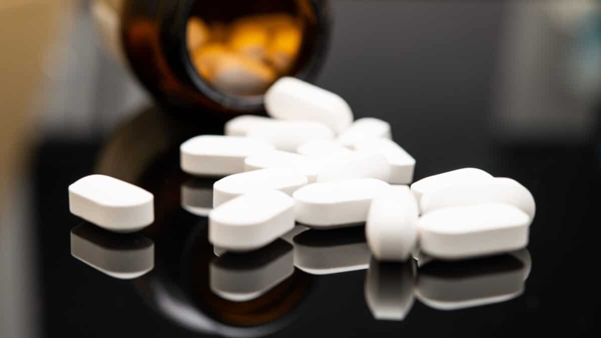 A close-up view of white, oval-shaped opioid tablets spilled from a dark prescription bottle onto a reflective black surface, highlighting pharmaceutical issues.