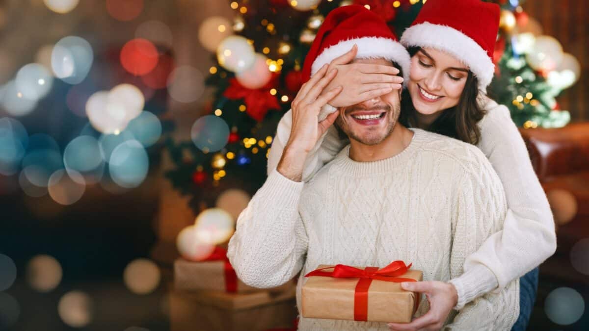 Loving young woman giving surprise gift to her husband, closing his eyes, celebrating Christmas at home